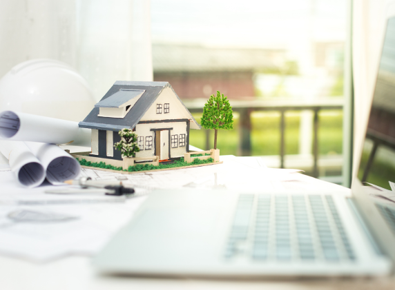 A miniature model of a house placed on a desk surrounded by architectural plans, a laptop, and a hard hat, suggesting a residential construction project.