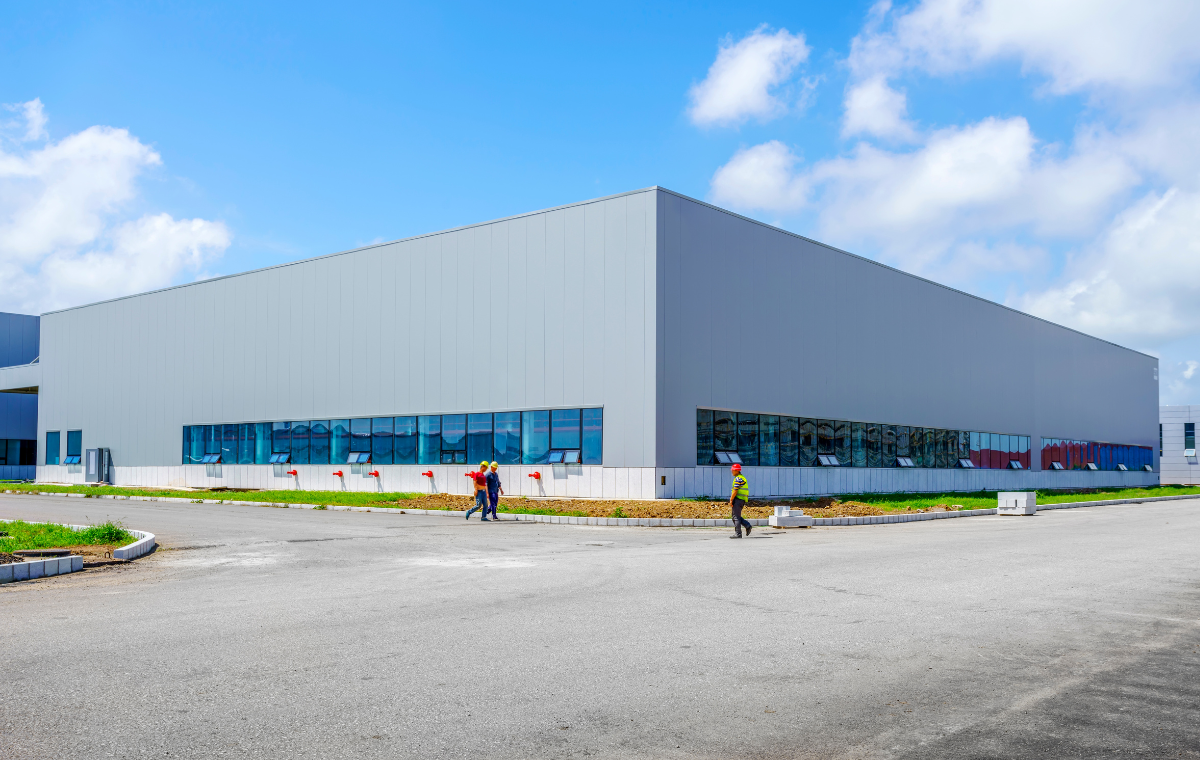 A large, modern commercial warehouse with a gray exterior and multiple windows along the side. Two workers in safety gear are walking on the paved area in front