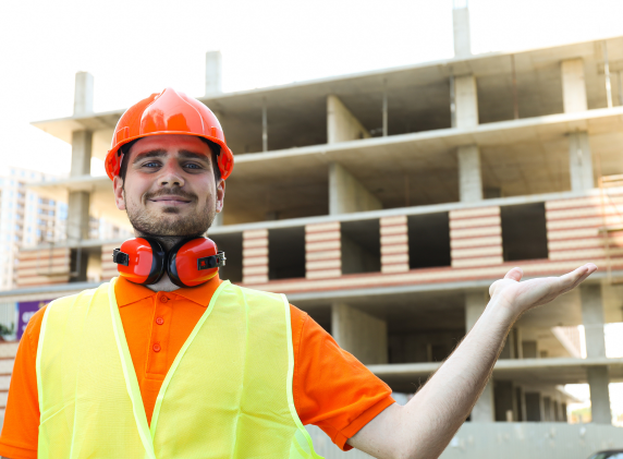 A construction worker in an orange hard hat and safety vest gestures toward a multi-story commercial building under construction.