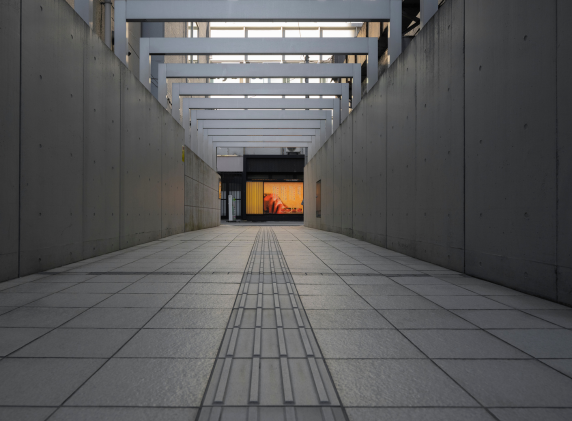 A modern industrial corridor with tiled flooring, concrete walls, and overhead beams leading to a brightly lit display