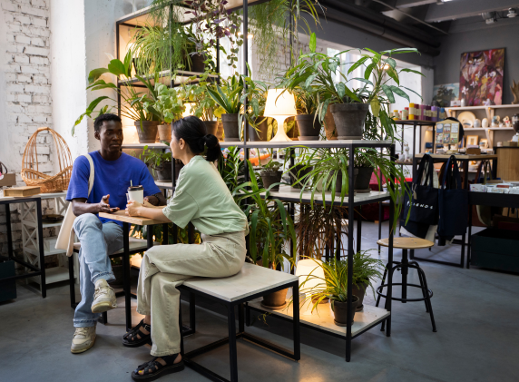 Two people sitting at a table in a modern, plant-filled interior space, engaged in conversation
