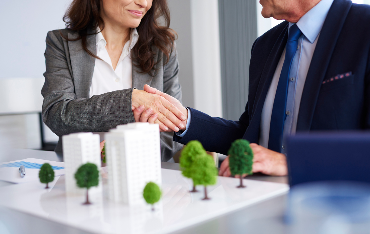 Two business professionals shaking hands over a table with architectural models and trees, symbolizing a successful agreement or partnership in a turnkey project