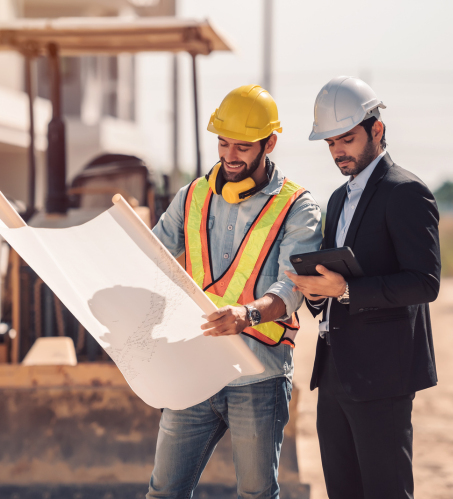 Two construction professionals wearing safety helmets and vests review a large blueprint at a construction site.