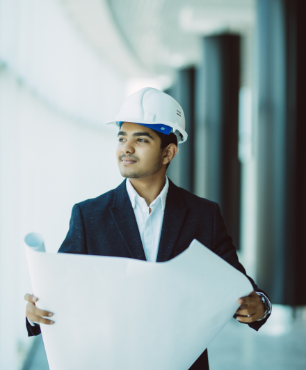 A person wearing a white hard hat and a dark suit is holding and examining a large blueprint in a modern, well-lit building.